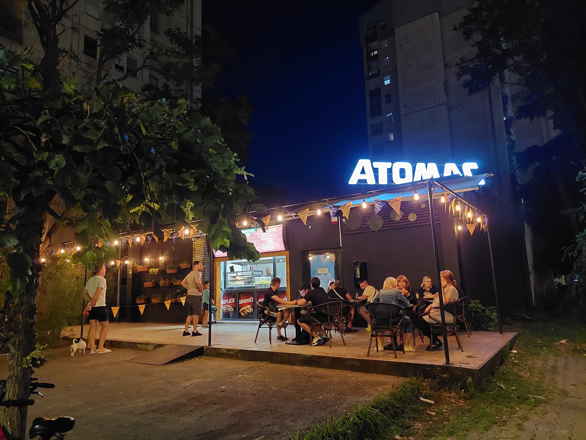 The Atomac kiosk at night: the lit sign, string lights and guests on the terrace